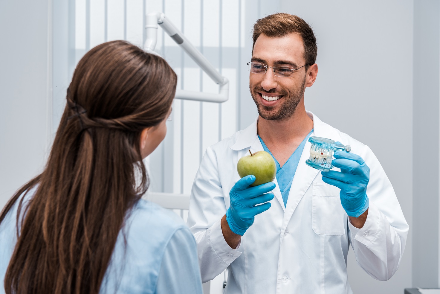 Happy dentist in glasses holding tooth model near patient